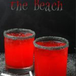 Two cocktail glasses with dark grey rims and orange liquid under the words Hex on the Beach